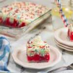 A slice of red white and blue poke cake topped with whipped cream and sprinkles sits on a plate, with the rest of the festive cake in a glass dish behind it. A blue checkered napkin and a glass of lemonade are nearby.