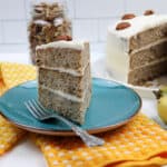Slice of Hummingbird Cake on a blue plate with fork beside the whole cake and jar of pecans.