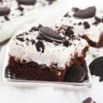 A close-up of a square piece of chocolate cake topped with creamy white frosting, crushed Oreo cookies, and an Oreo on top, perfect for anyone searching for the ultimate Oreo poke cake recipe. More Oreos and another cake slice appear in the background.