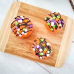 Three orange frosted treats topped with black drizzle and colorful sprinkles sit on a wooden cutting board, perfect for a Halloween Dessert Table. Black straws with orange dots are placed nearby on a light background.
