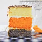 A slice of pumpkin spice latte cake with cream cheese frosting features white, orange, and gray layers on a checkered plate, with a cake stand and another slice in the background. Orange cake crumbs are scattered on the plate.