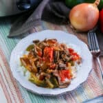 A plate of Slow Cooker Pepper Steak with shredded beef, green and red bell peppers, and savory sauce sits on a striped placemat. A fork, onion, fresh bell peppers, and a slow cooker are nearby.
