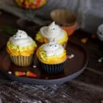 Three Halloween-themed cupcakes with yellow and orange frosting, topped with ghost-shaped meringues and candy eyes, sit on a dark plate—perfect for a Halloween Dessert Table. Candy corn pieces and cupcake wrappers are scattered around.