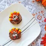 Two cupcakes decorated as turkeys with candy corn feathers and candy eyes sit on a white plate alongside festive thanksgiving brownies. The plate rests on a patterned tablecloth with decorative leaves and a small pumpkin nearby.