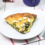 A breakfast setup with a slice of frittata on a plate, a mug of coffee, and the skillet of frittata in the background.