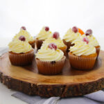 Six cranberry orange cupcakes with swirled white frosting and sugared cranberries on top are arranged on a round wooden platter, which sits on a gray cloth. The background is white and bright.