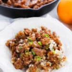 A white plate with Panda Express Orange Chicken Recipe and rice, garnished with sliced green onions. In the background, a skillet with more orange chicken and a whole orange are visible on a light-colored cloth.
