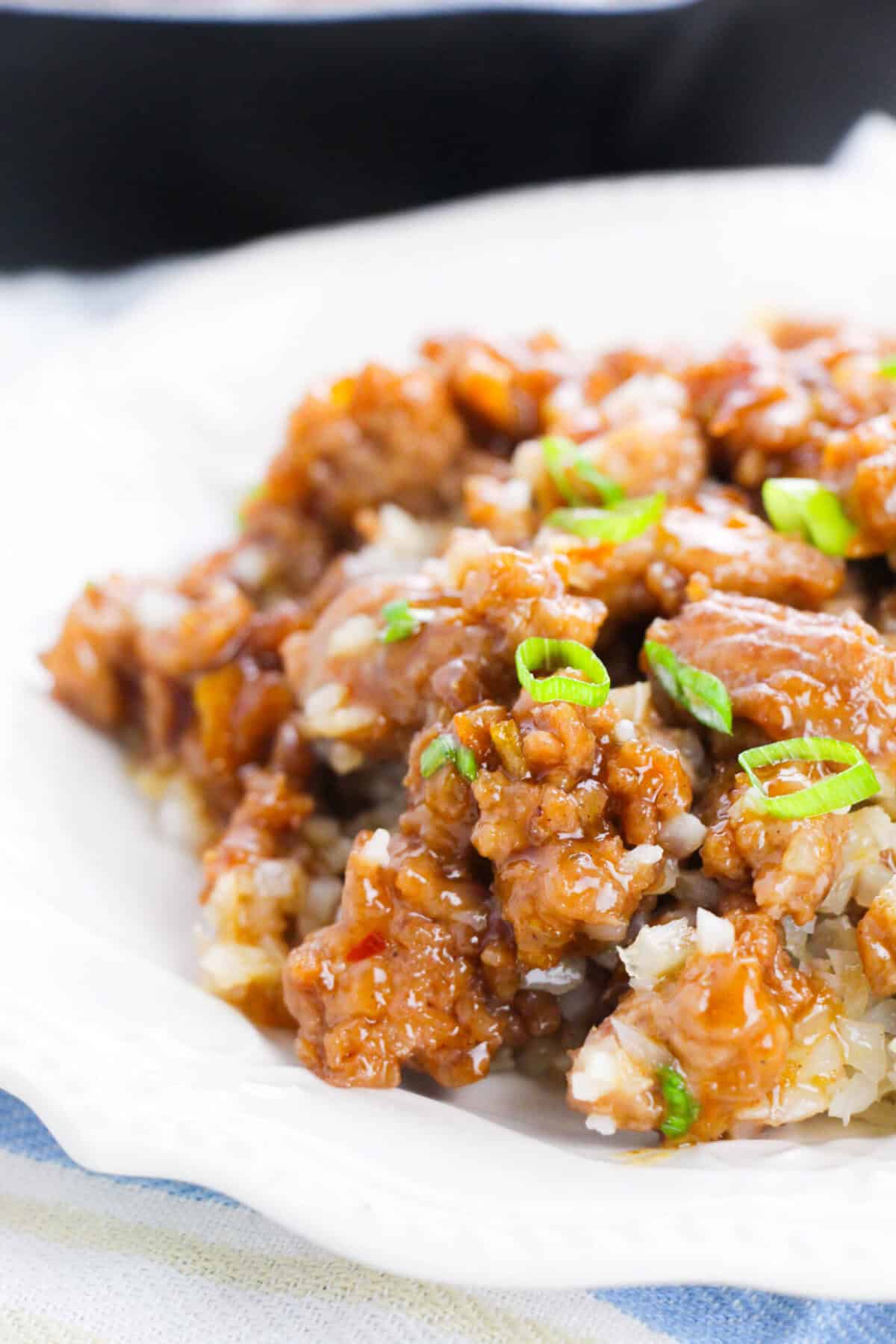 A close-up of a plate of saucy, breaded chicken pieces topped with chopped green onions, served over a bed of rice on a white dish.