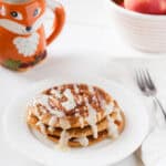 A stack of pumpkin cinnamon roll pancakes drizzled with icing sits on a white plate next to a fork and napkin, with a fox-themed mug and a bowl of apples and lemons in the background.