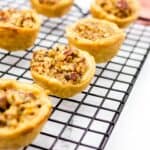 Mini pecan pies cooling on a black wire rack, with golden, flaky crusts and a rich pecan filling. Perfect for christmas dessert ideas, these treats sit against a white background with a hint of red-striped cloth.