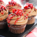 A tray of frosted cupcakes with cherry topping on a checkered cloth.