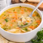 A bowl of chicken soup with carrots, celery, and herbs, served with saltine crackers and garnished with fresh parsley—one of the best comfort food ideas. A spoon rests in the soup; salt, pepper shakers, and bread are in the background.