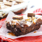 A slice of Snickers poke cake on a red plate in front of the full cake in a glass dish.