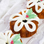 Mini bundt cakes decorated with white icing in a bow shape and topped with green holly leaf and red berry decorations, arranged on an ornate white platter—perfect as cute Christmas desserts for your festive table.