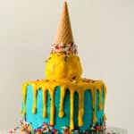 A blue frosted melted ice cream cone cake, decorated with colorful sprinkles, yellow icing dripping down the sides, and an upside-down cone on top coated in yellow icing and sprinkles—perfect for any melted ice cream cone cake recipe.