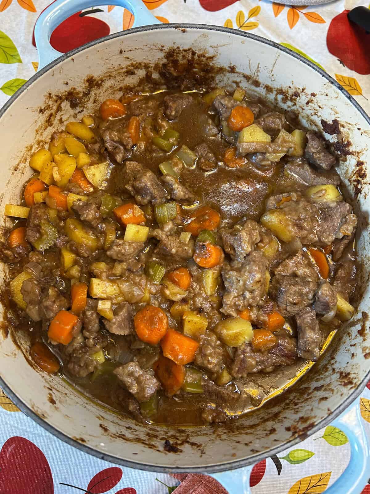 A pot of Dutch Oven Beef Stew with chunks of beef, carrots, potatoes, and celery in a thick brown broth sits on a table with a colorful leaf-patterned tablecloth.