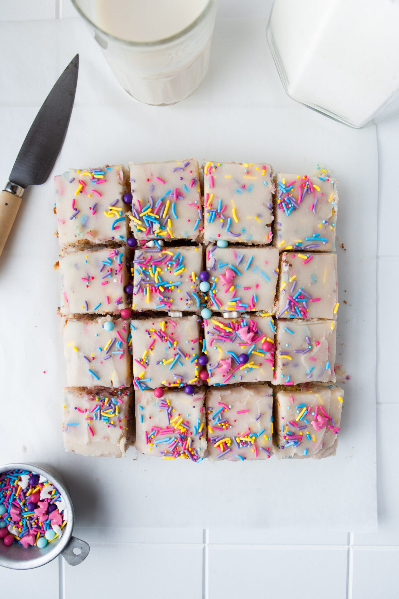 Sliced funfetti sheet cake topped with colorful sprinkles, with a glass of milk and a container of sugar in the background.