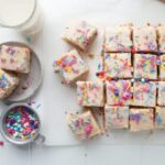 Sliced funfetti sheet cake with colorful sprinkles, surrounded by milk, a plate of cake, and a sprinkle bowl.