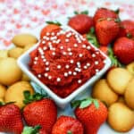 A white bowl filled with creamy red velvet dip topped with white sprinkles, surrounded by fresh strawberries and vanilla wafer cookies on a heart-patterned placemat.