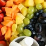 A plate arranged with sliced strawberries, cantaloupe, pineapple, green grapes, and blueberries forms a vibrant fruit rainbow. Marshmallows and gold-wrapped candies sit in bowls nearby, accented by green shamrock decorations.
