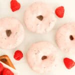 Four donuts with pink glaze, surrounded by fresh raspberries.