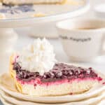 A slice of berry pie with a creamy filling, topped with whipped cream, sits on stacked plates. The rest of the pie, inspired by classic blueberry pie recipes, is on a cake stand in the background, next to a cup of coffee.