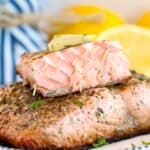 A close-up of an Olive Garden Herb Grilled Salmon fillet topped with a pat of butter, served on a decorative plate with sliced lemons and a blue-striped cloth in the blurred background.