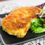 An Old Fashioned Breaded Pork Chop, golden-brown and crisp, is served on a black plate beside a small mixed green salad, all arranged on a printed cloth.