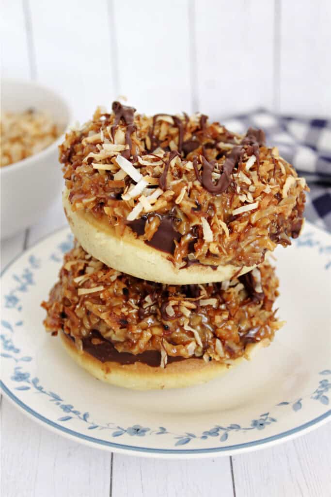 Two donuts topped with chocolate, caramel, and toasted shredded coconut are stacked on a floral plate, with a bowl of extra coconut and a checkered cloth in the background.