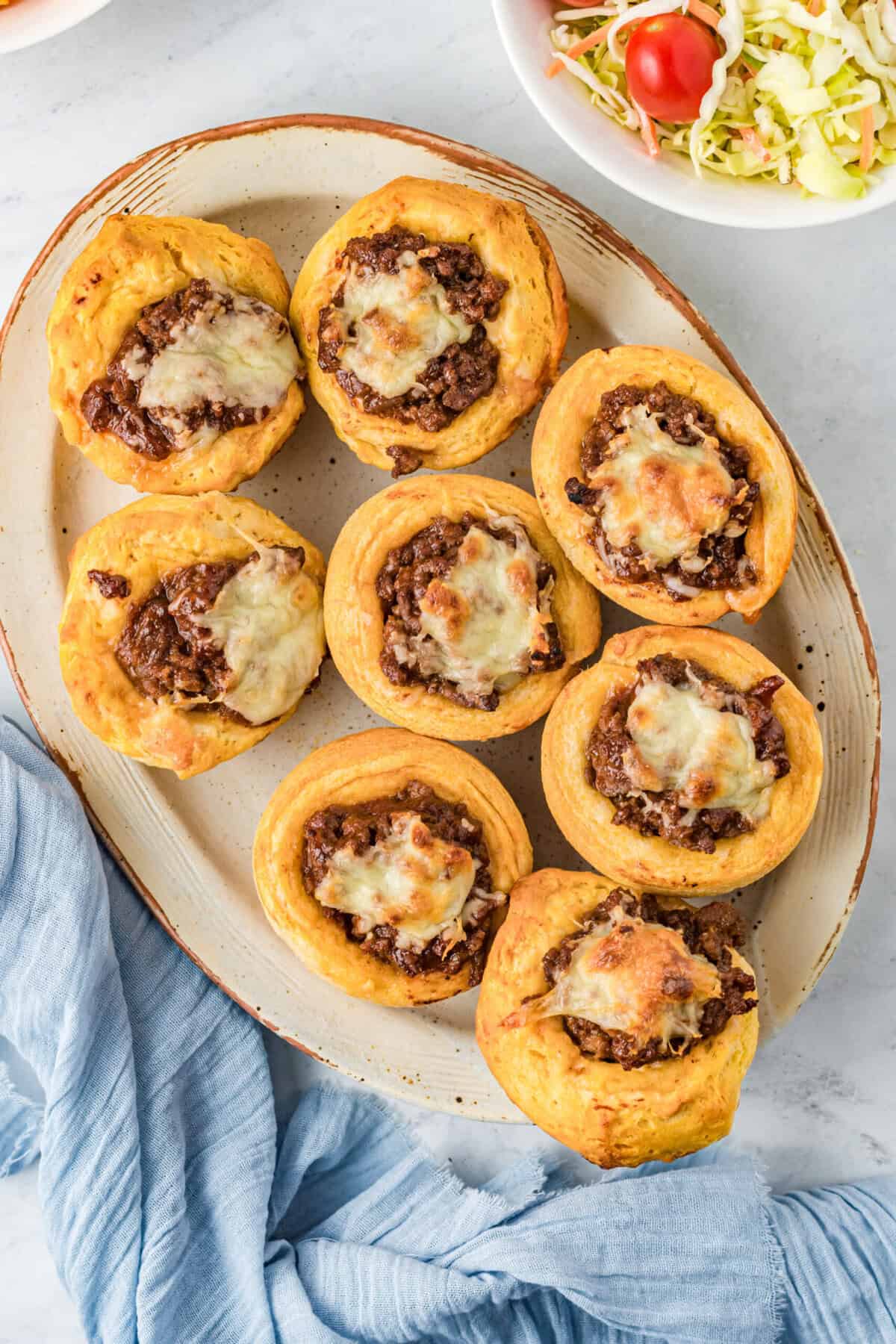 A platter of eight baked biscuit cups filled with seasoned ground beef and melted cheese, placed on a light surface with a blue cloth nearby and a bowl of fresh salad in the corner.