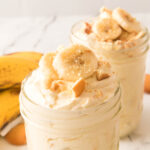 Two mason jars filled with creamy banana pudding, topped with banana slices and crumbled vanilla wafers—an irresistible treat that’s perfect for fans of ripe banana recipes. Bananas and whole vanilla wafers sit in the background on a white marble surface.