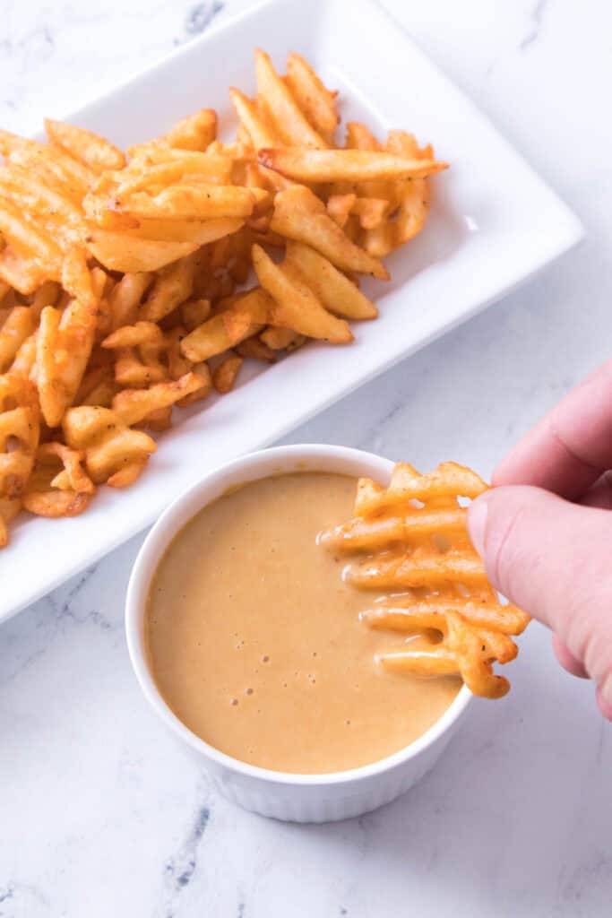 A hand dips a seasoned waffle fry into a small white bowl of creamy sauce, with a plate of more waffle fries in the background on a white marble surface.