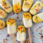 Ten white chocolate-covered cakesicles are arranged in a circle, some topped with yellow drizzle and colorful sprinkles, with extra sprinkles scattered on a light gray surface.