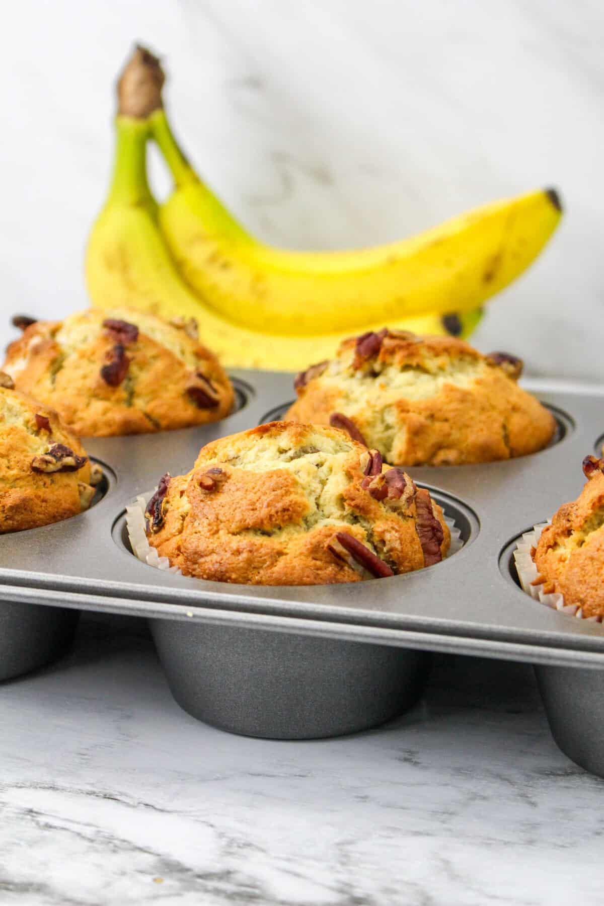 A muffin tin filled with freshly baked banana nut muffins sits on a marble surface, with two ripe bananas in the background, highlighting the delicious flavor of homemade banana nut muffins.