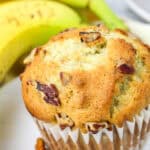 A close-up of a banana nut muffin in a paper liner, topped with chopped nuts. Two ripe bananas and scattered pecan pieces are visible in the background, capturing the classic appeal of freshly baked banana nut muffins.