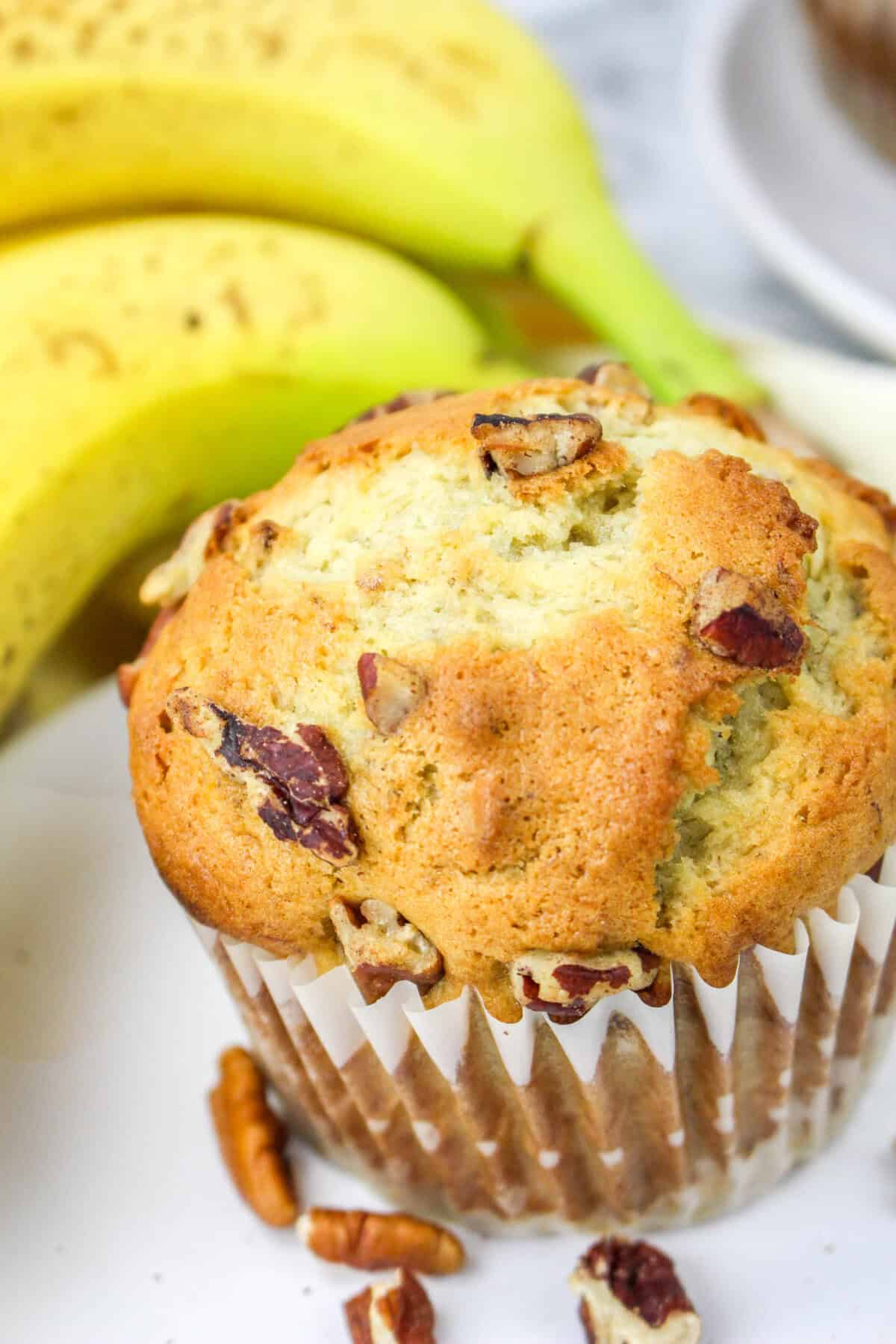 A close-up of a banana nut muffin in a paper liner, topped with chopped nuts. Two ripe bananas and scattered pecan pieces are visible in the background, capturing the classic appeal of freshly baked banana nut muffins.