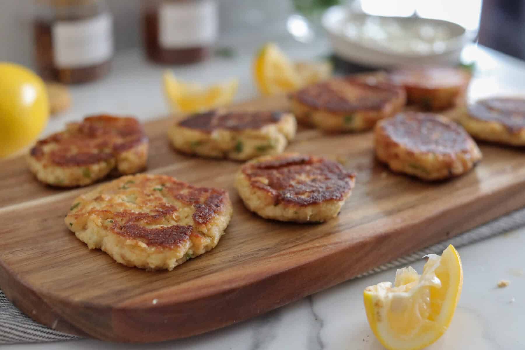 Golden brown crab cakes are arranged on a wooden serving board, with lemon wedges scattered around. The background shows blurred jars and a bowl, suggesting a casual, homemade meal setting.