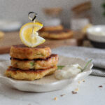 Three golden-brown patties stacked on a plate, garnished with a lemon wedge and a dollop of creamy sauce—perfect Memorial Day finger foods. In the background are more patties, a bowl of sauce, and blurred kitchen items.