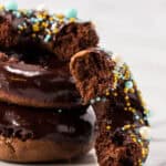 Three chocolate-glazed homemade donuts are stacked, topped with colorful blue, yellow, and white sprinkles. The top donut has a large bite taken out, revealing its soft, rich interior. The background is a white marble surface.