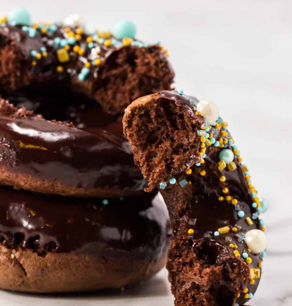 Three chocolate-glazed homemade donuts are stacked, topped with colorful blue, yellow, and white sprinkles. The top donut has a large bite taken out, revealing its soft, rich interior. The background is a white marble surface.