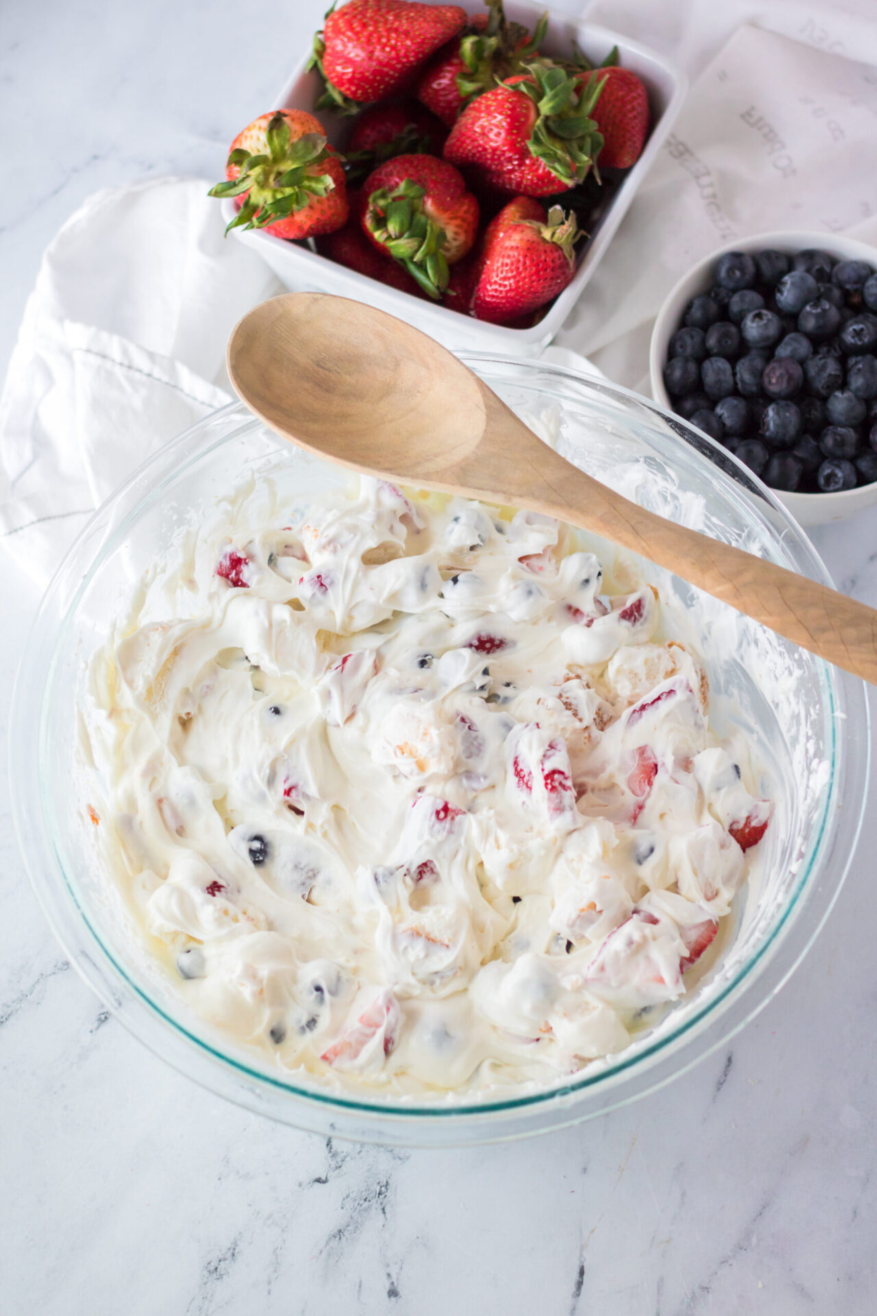 A glass bowl filled with creamy fruit salad containing strawberries and blueberries, with a wooden spoon resting on the bowl. Fresh strawberries and blueberries in separate bowls are in the background.