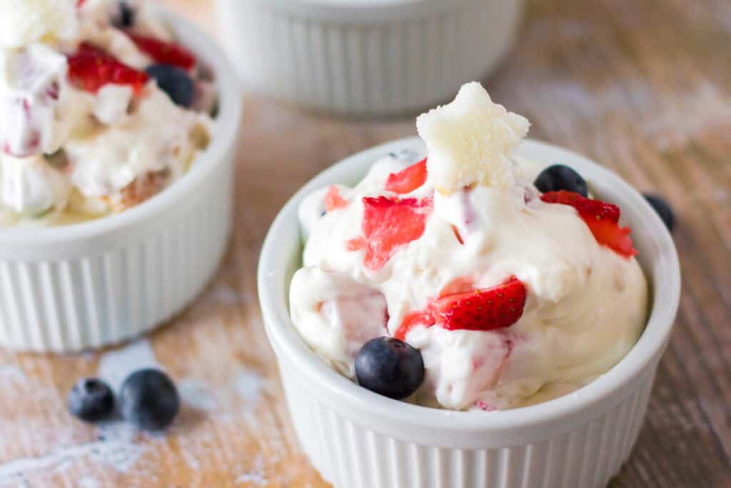 A white ramekin filled with creamy Very Berry Cheesecake Salad, topped with sliced strawberries, blueberries, and a small star-shaped garnish. Another ramekin with a similar dessert is visible in the background.