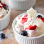 A white ramekin filled with creamy Very Berry Cheesecake Salad, topped with sliced strawberries, blueberries, and a small star-shaped garnish. Another ramekin with a similar dessert is visible in the background.