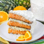 A slice of layered sunset cake, combining orange and coconut with white frosting and coconut flakes, is garnished with orange slices on a white plate, while a whole orange, pineapple, and the full cake rest on a colorful striped cloth.