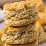 A close-up of freshly baked, golden-brown Pancake Mix Biscuits stacked on top of each other, showing their flaky layers and soft texture. More biscuits are visible in the blurred background.