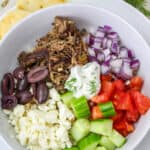 A white bowl filled with shredded meat, chopped red onion, diced tomatoes, sliced cucumber, kalamata olives, crumbled feta cheese, and tzatziki sauce—an irresistible dinner idea for summer nights. The bowl sits on a white plate with a fork beside it.