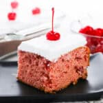 A slice of cherry cake with white frosting and a cherry on top on a black plate.