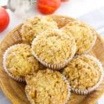 A basket filled with six apple muffins topped with crumbly streusel, surrounded by fresh apples, a stick of butter, and a measuring cup of flour—perfect for cozy fall breakfast recipes.