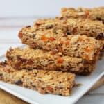 A stack of homemade granola bars, inspired by a classic muesli bar recipe, with oats, dried fruit, and nuts is arranged on a white rectangular plate atop a wooden board.