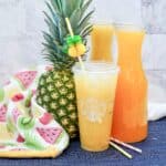 A Starbucks cup filled with yellow pineapple drink sits in front of a whole pineapple, two glass carafes of juice, and a colorful fruit-patterned towel—perfect inspiration for your next starbucks drink recipes. Two striped straws and a pineapple stirrer are nearby.